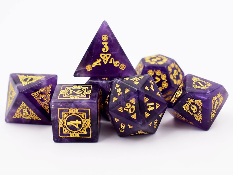 May include: Set of six purple and gold dice with Celtic knot designs. The dice are made of amethyst and have gold numbers and symbols.