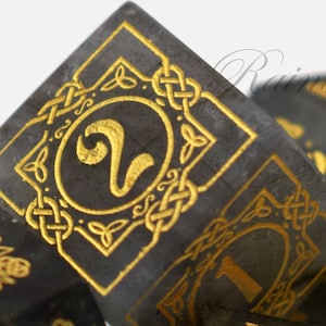 May include: A set of black and gold dice with Celtic knotwork designs. The dice are made of a dark, opaque material and have gold-colored numbers and designs. The dice are numbered 1 through 6.