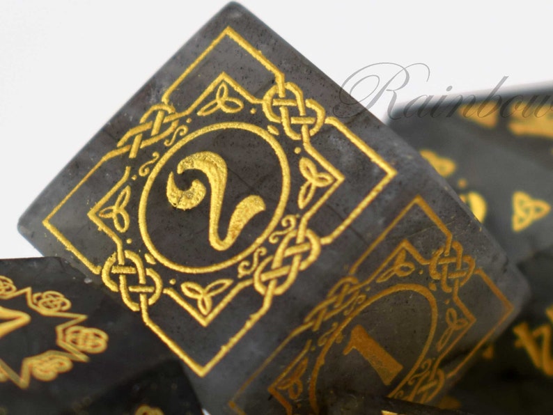 May include: A set of black and gold dice with Celtic knotwork designs. The dice are made of a dark, opaque material and have gold-colored numbers and designs. The dice are numbered 1 through 6.