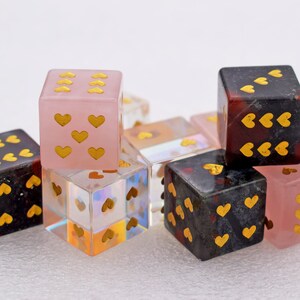 K&ouml;nnte beinhalten: Ein Set aus sechs herzf&ouml;rmigen W&uuml;rfeln in verschiedenen Farben, darunter rosa, schwarz und klar. Die W&uuml;rfel sind aus Stein und haben goldfarbene Herzen auf jeder Seite.