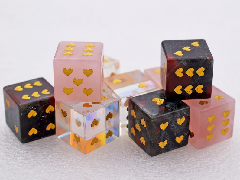 K&ouml;nnte beinhalten: Ein Set aus sechs herzf&ouml;rmigen W&uuml;rfeln in verschiedenen Farben, darunter rosa, schwarz und klar. Die W&uuml;rfel sind aus Stein und haben goldfarbene Herzen auf jeder Seite.