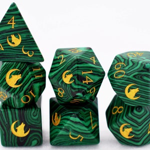Puede incluir: Un juego de dados poli&eacute;dricos con un patr&oacute;n verde malaquita y negro. Los dados tienen n&uacute;meros dorados y un s&iacute;mbolo dorado de lobo y luna creciente. El juego incluye un dado de cuatro caras, un dado de seis caras, un dado de ocho caras, un dado de diez caras, un dado de doce caras, un dado de veinte caras y un dado de porcentaje.
