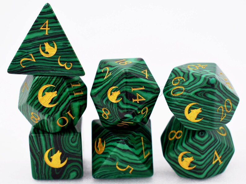 Puede incluir: Un juego de dados poli&eacute;dricos con un patr&oacute;n verde malaquita y negro. Los dados tienen n&uacute;meros dorados y un s&iacute;mbolo dorado de lobo y luna creciente. El juego incluye un dado de cuatro caras, un dado de seis caras, un dado de ocho caras, un dado de diez caras, un dado de doce caras, un dado de veinte caras y un dado de porcentaje.