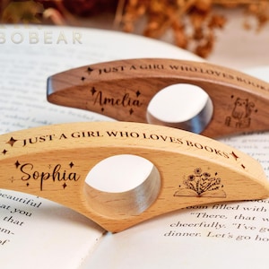 Op de afbeelding: Twee houten duim-paginahouders met een gat in het midden. De voorste is van licht hout, gegraveerd met "JUST A GIRL WHO LOVES BOOKS", "Sophia" en een bloemenboek-icoon. De achterste is van donker hout, gegraveerd met "JUST A GIRL WHO LOVES BOOKS", "Amelia" en een kat-icoon.