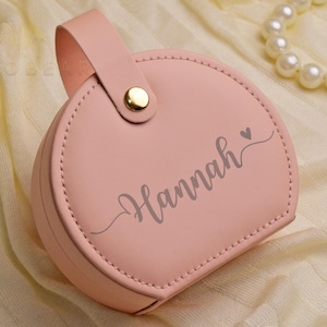 K&ouml;nnte beinhalten: Eine hellrosa, runde Schmuckschatulle mit Griff und goldenem Verschluss. Der Name "Hannah" ist auf der Vorderseite geschrieben. Eine Perlenkette ist im Hintergrund. Das Etui ist wahrscheinlich aus Leder oder einem &auml;hnlichen Material.