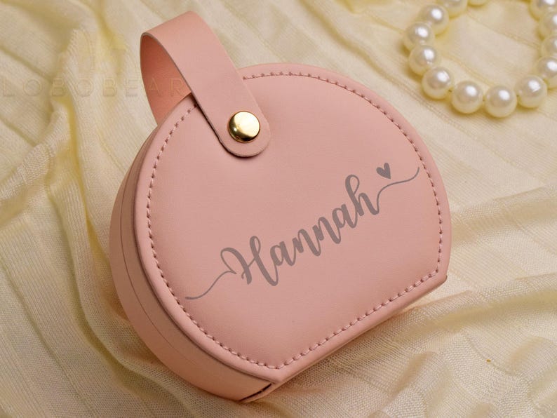 K&ouml;nnte beinhalten: Eine hellrosa, runde Schmuckschatulle mit Griff und goldenem Verschluss. Der Name "Hannah" ist auf der Vorderseite geschrieben. Eine Perlenkette ist im Hintergrund. Das Etui ist wahrscheinlich aus Leder oder einem &auml;hnlichen Material.
