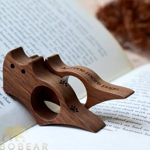 Puede incluir: Dos sujetap&aacute;ginas de madera con forma de gato con huellas de patas y el texto "Just one more page..." grabado. Los accesorios para libros son de madera oscura y est&aacute;n sobre un libro abierto.