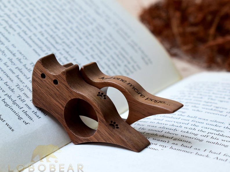 Puede incluir: Dos sujetap&aacute;ginas de madera con forma de gato con huellas de patas y el texto "Just one more page..." grabado. Los accesorios para libros son de madera oscura y est&aacute;n sobre un libro abierto.