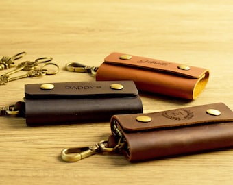 May include: Three leather key holders in shades of brown, with brass hardware. One is dark brown with "DADDY" engraved, another is tan with "Gabrielle" engraved, and the third is brown with an "M" and a laurel wreath.