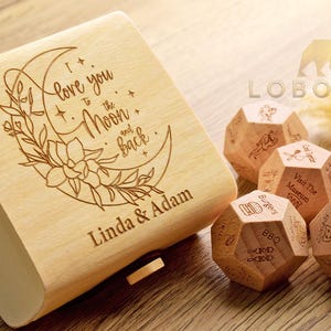 Könnte beinhalten: Eine helle Holzschachtel, graviert mit einer Mondsichel, floralen Elementen und dem Text "I love you to the Moon and back" sowie den Namen "Linda & Adam". Drei hölzerne Dodekaeder-Würfel mit verschiedenen eingravierten Symbolen und Texten.