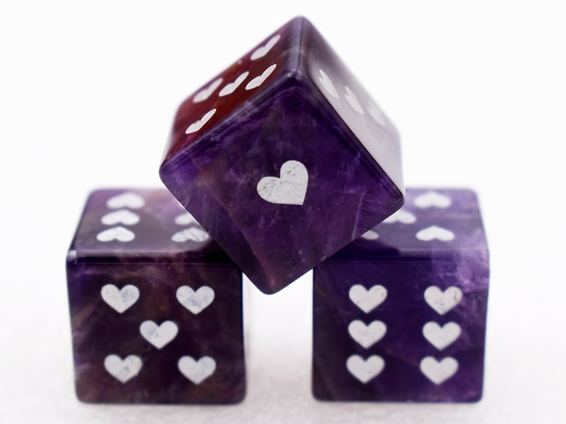 K&ouml;nnte beinhalten: Drei violette Amethyst-W&uuml;rfel mit wei&szlig;en Herzsymbolen auf jeder Seite. Die W&uuml;rfel sind &uuml;bereinander gestapelt.