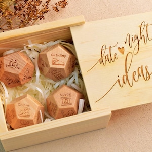 Könnte beinhalten: Eine helle Holzschachtel mit einem Schiebedeckel, der mit dem Text date night ideas und einem kleinen Herz graviert ist. Im Inneren befinden sich drei polyedrische Holzwürfel mit verschiedenen eingravierten Illustrationen und Beschriftungen wie Chicken und Go To Sleep.