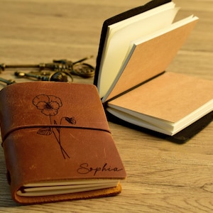 以下が含まれることがあります： 木製の表面に、閉じた状態と開いた状態の2つの革製ジャーナルがあります。 閉じたジャーナルは茶色で、花の模様と「Sophia」という名前が刻まれています。 開いたジャーナルには、ベージュと白のページがあります。