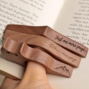 May include: Three brown wooden page holders with a thumb hole. Two of the holders are engraved with the text "Just one more page..." in a cursive font, while the third holder features a simple line drawing of a mountain range on its surface.