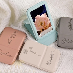 Op de afbeelding: Een verzameling gepersonaliseerde compacte spiegels in verschillende kleuren. Eén is open en toont een bloemenafbeelding. Elke compact heeft een bloemenontwerp en een naam: Sophia, Hannah en Gabrielle. De open spiegel is lichtblauw.