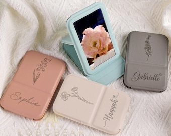 Può includere: Una collezione di specchietti compatti personalizzati in vari colori. Uno è aperto, mostrando l'immagine di un fiore. Ogni specchietto presenta un disegno floreale e un nome: Sophia, Hannah e Gabrielle. Lo specchietto aperto è azzurro.