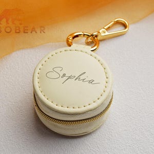 May include: A round, cream-colored, zippered jewelry case featuring the name Sophia in elegant script on the top lid. The case includes a gold-toned metal lobster clasp attachment at the top for easy hanging or securing to other items.