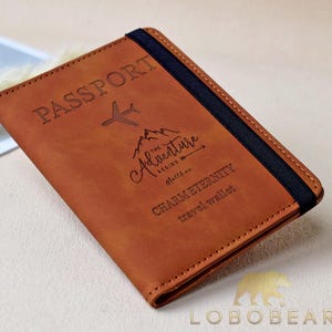 Könnte beinhalten: Braunes Leder-Reisepass-Etui mit dem geprägten Wort „PASSPORT“ oben. Es zeigt eine Flugzeug-Grafik und den Satz „THE ADVENTURE BEGINS“ mit „CHARMETERNITY travel wallet“ darunter. Ein schwarzes Gummiband sichert das Portemonnaie.