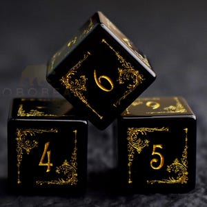 Peut inclure: Trois d&eacute;s noirs avec des chiffres dor&eacute;s et des motifs d&eacute;coratifs. Les d&eacute;s sont empil&eacute;s, le d&eacute; du dessus affichant le chiffre 6. Les autres chiffres visibles sont 4 et 5. Les d&eacute;s ont une finition brillante.