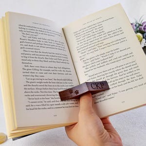 May include: A wooden bookmark with a paw print design and the letters "CKP" engraved on it. The bookmark is placed on an open book with text about dwarves and a giant.