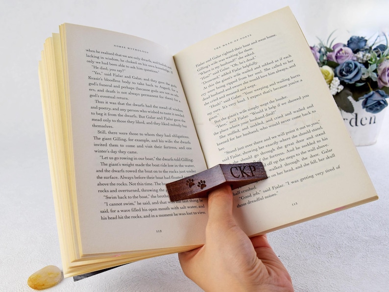 May include: A wooden bookmark with a paw print design and the letters "CKP" engraved on it. The bookmark is placed on an open book with text about dwarves and a giant.