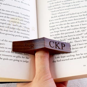 May include: A wooden bookmark with a paw print design and the initials "CKP" engraved on it. The bookmark is being used to hold open a book.