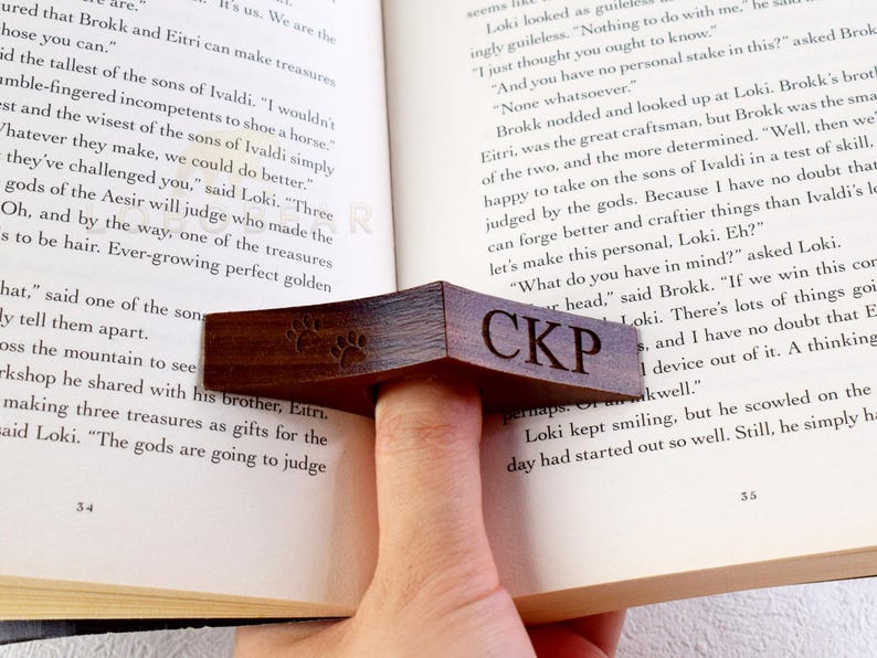 May include: A wooden bookmark with a paw print design and the initials "CKP" engraved on it. The bookmark is being used to hold open a book.