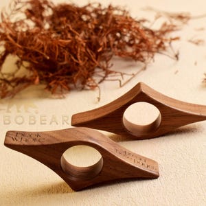 May include: Two wooden book holders with a circular opening and angled design. One has the text "Book whore" and the other reads "spread those pages". The holders are made of a dark wood and are on a light beige surface.