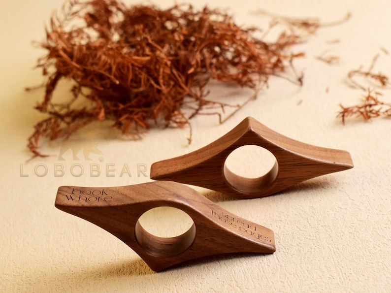 May include: Two wooden book holders with a circular opening and angled design. One has the text "Book whore" and the other reads "spread those pages". The holders are made of a dark wood and are on a light beige surface.