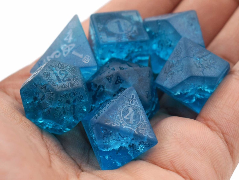 May include: A set of six blue dice with intricate designs and numbers etched into the surface. The dice are made of a translucent material, giving them a frosted appearance.