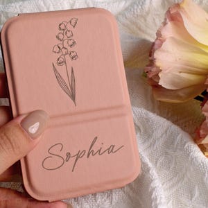Könnte beinhalten: Eine hellrosa Handyhülle mit floralem Design und dem Schriftzug "Sophia". Die Hülle zeigt eine Maiglöckchen-Illustration. Der Hintergrund umfasst einen weißen Stoff und eine rosa-gelbe Blume.