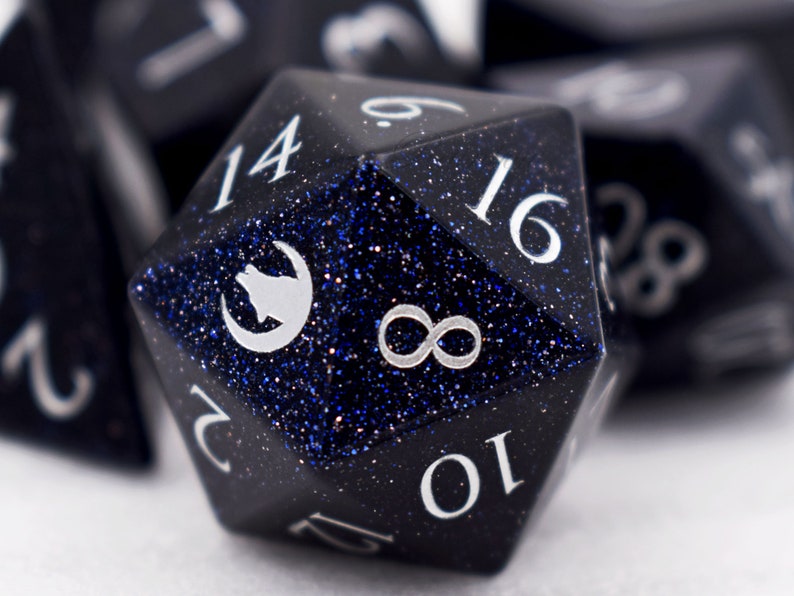 Puede incluir: Primer plano de un dado poli&eacute;drico con una superficie azul oscuro y brillante. El dado tiene n&uacute;meros blancos y un s&iacute;mbolo blanco de lobo y luna creciente. Otros dados son visibles en el fondo, fuera de foco. El dado es para juegos de mesa.