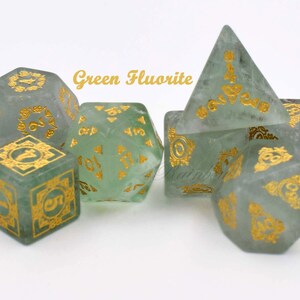 May include: Set of six green fluorite dice with gold accents. The dice are in various shapes, including a pyramid, a cube, and a d20. The dice are on a white surface.