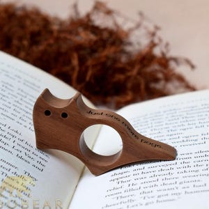 Puede incluir: Un sujetap&aacute;ginas de madera con forma de gato con el texto "Just one more page..." grabado. El sujetap&aacute;ginas est&aacute; apoyado en un libro abierto, con un fondo borroso de material vegetal seco.