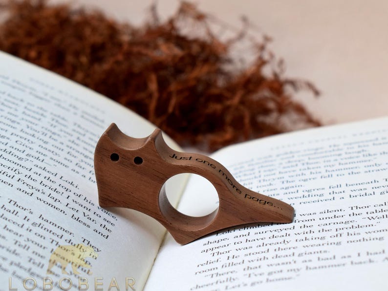 Puede incluir: Un sujetap&aacute;ginas de madera con forma de gato con el texto "Just one more page..." grabado. El sujetap&aacute;ginas est&aacute; apoyado en un libro abierto, con un fondo borroso de material vegetal seco.