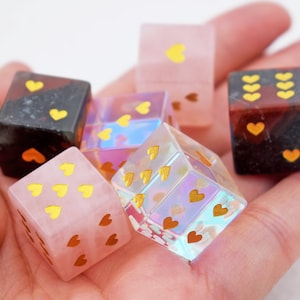 K&ouml;nnte beinhalten: Sechs herzf&ouml;rmige W&uuml;rfel in verschiedenen Farben und Materialien, darunter rosa, schwarz, klar und schillernd. Die W&uuml;rfel sind mit goldenen Herzen verziert.