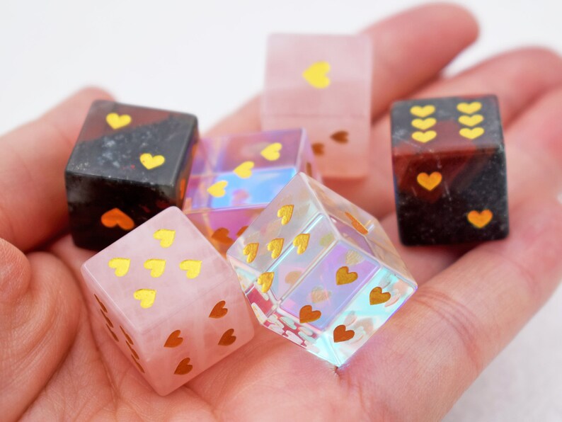 K&ouml;nnte beinhalten: Sechs herzf&ouml;rmige W&uuml;rfel in verschiedenen Farben und Materialien, darunter rosa, schwarz, klar und schillernd. Die W&uuml;rfel sind mit goldenen Herzen verziert.
