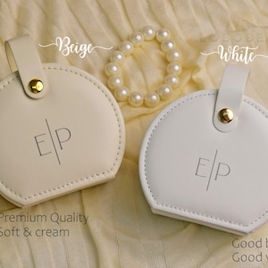 K&ouml;nnte beinhalten: Zwei runde, cremefarbene Etuis mit Griff und goldenem Knopfverschluss, neben einem Perlenarmband. Die Etuis tragen die Initialen "E/P". Eines ist mit "Beige" und das andere mit "White" beschriftet. Der Text "Premium Quality Soft & cream" ist sichtbar.
