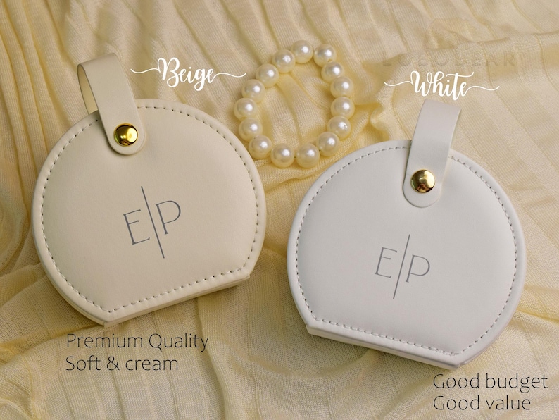 K&ouml;nnte beinhalten: Zwei runde, cremefarbene Etuis mit Griff und goldenem Knopfverschluss, neben einem Perlenarmband. Die Etuis tragen die Initialen "E/P". Eines ist mit "Beige" und das andere mit "White" beschriftet. Der Text "Premium Quality Soft & cream" ist sichtbar.