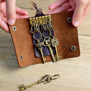 Könnte beinhalten: Ein brauner Leder-Schlüsselhalter mit mehreren Schlüsseln im antiken Stil. Die Schlüssel sind aus Messing und weisen verzierte Designs auf. Ein schlüssel in Form eines Seepferdchens liegt separat. Der Schlüsselhalter ist geöffnet und zeigt die Schlüssel.
