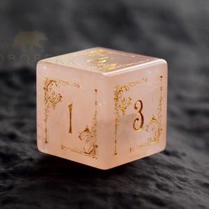 Peut inclure: Un d&eacute; rose clair de forme carr&eacute;e, orn&eacute; de chiffres dor&eacute;s et de motifs filigran&eacute;s. Le d&eacute; affiche les chiffres 1 et 3 sur des faces adjacentes. Il semble &ecirc;tre fait d'un mat&eacute;riau translucide, possiblement de la pierre ou de la r&eacute;sine.