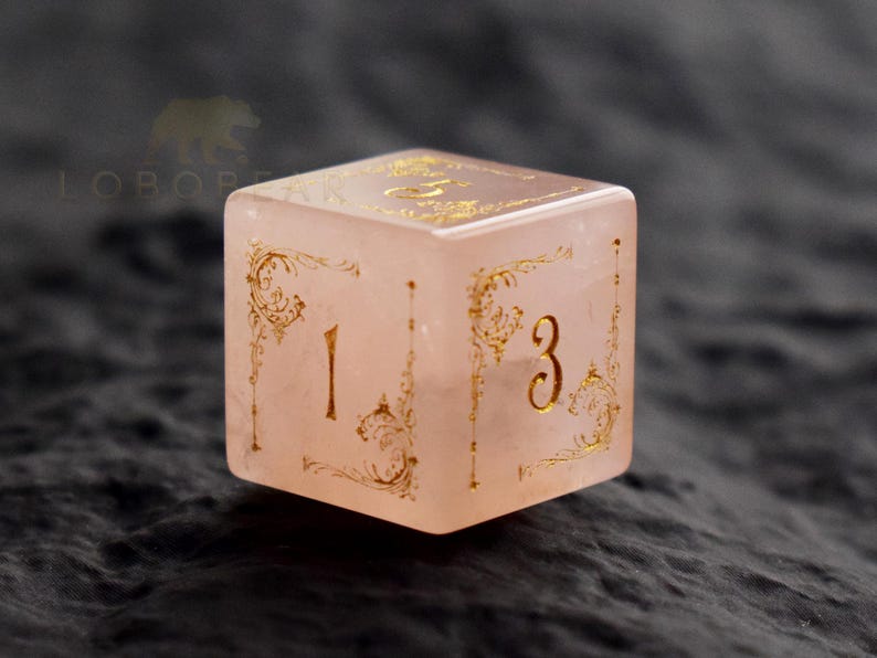 Peut inclure: Un d&eacute; rose clair de forme carr&eacute;e, orn&eacute; de chiffres dor&eacute;s et de motifs filigran&eacute;s. Le d&eacute; affiche les chiffres 1 et 3 sur des faces adjacentes. Il semble &ecirc;tre fait d'un mat&eacute;riau translucide, possiblement de la pierre ou de la r&eacute;sine.