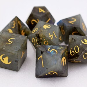 Puede incluir: Un conjunto de siete dados de piedras preciosas con n&uacute;meros dorados y una silueta de lobo. Los dados son una mezcla de gris y azul con un efecto brillante. Los dados son para juegos de mesa.