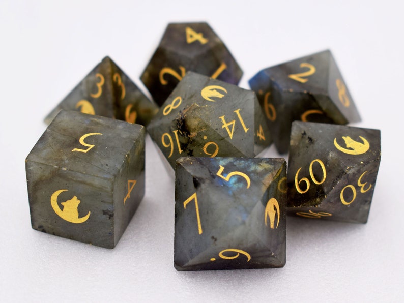 Puede incluir: Un conjunto de siete dados de piedras preciosas con n&uacute;meros dorados y una silueta de lobo. Los dados son una mezcla de gris y azul con un efecto brillante. Los dados son para juegos de mesa.