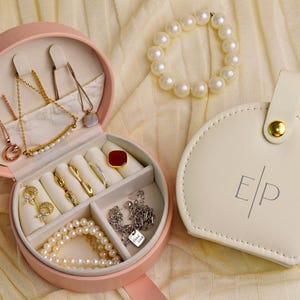 K&ouml;nnte beinhalten: Eine offene, rosa Schmuckschatulle mit Halsketten, Ringen und einem Perlenarmband. Ein wei&szlig;es, rundes Etui mit den Initialen "E/P" befindet sich neben der Schatulle. Die Schmuckschatulle ist mit verschiedenen Gold- und Silberschmuck gef&uuml;llt.