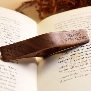May include: A wooden book page holder with the words "book whore" and "spread those pages" engraved on it. The holder is placed on an open book, with the pages visible. The wood has a rich, dark brown color.