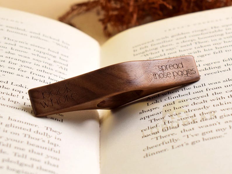 May include: A wooden book page holder with the words "book whore" and "spread those pages" engraved on it. The holder is placed on an open book, with the pages visible. The wood has a rich, dark brown color.