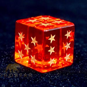 Puede incluir: Un dado rojo transparente con diseños de estrellas doradas en cada cara. El dado está iluminado, creando un efecto brillante. La imagen está sobre un fondo oscuro y texturizado, resaltando los detalles del dado.
