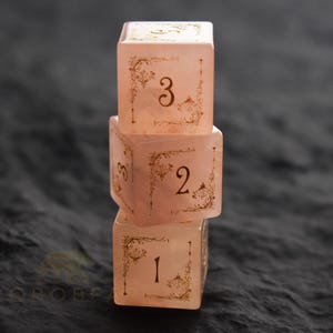 Peut inclure: Trois d&eacute;s roses translucides empil&eacute;s, avec des chiffres dor&eacute;s et des motifs d&eacute;coratifs aux coins. Les d&eacute;s sont num&eacute;rot&eacute;s 1, 2 et 3, sur un fond sombre et textur&eacute;. Les d&eacute;s semblent &ecirc;tre en r&eacute;sine.
