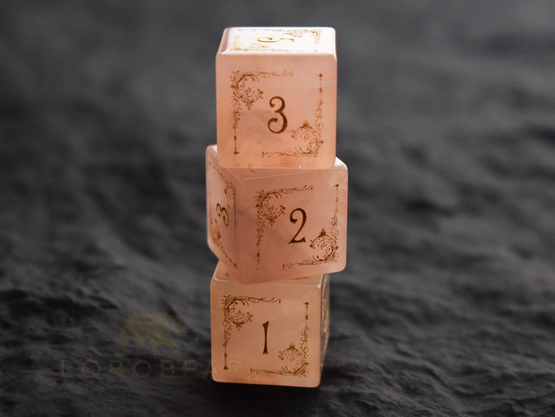 Peut inclure: Trois d&eacute;s roses translucides empil&eacute;s, avec des chiffres dor&eacute;s et des motifs d&eacute;coratifs aux coins. Les d&eacute;s sont num&eacute;rot&eacute;s 1, 2 et 3, sur un fond sombre et textur&eacute;. Les d&eacute;s semblent &ecirc;tre en r&eacute;sine.